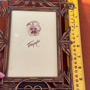 May include: A rectangular bronze-coloured metal picture frame with ornate leaf-like scrollwork detailing. A yellow ruler placed along the side indicates a height of approximately 25.4 cm. The frame contains a display card with a pansy illustration.