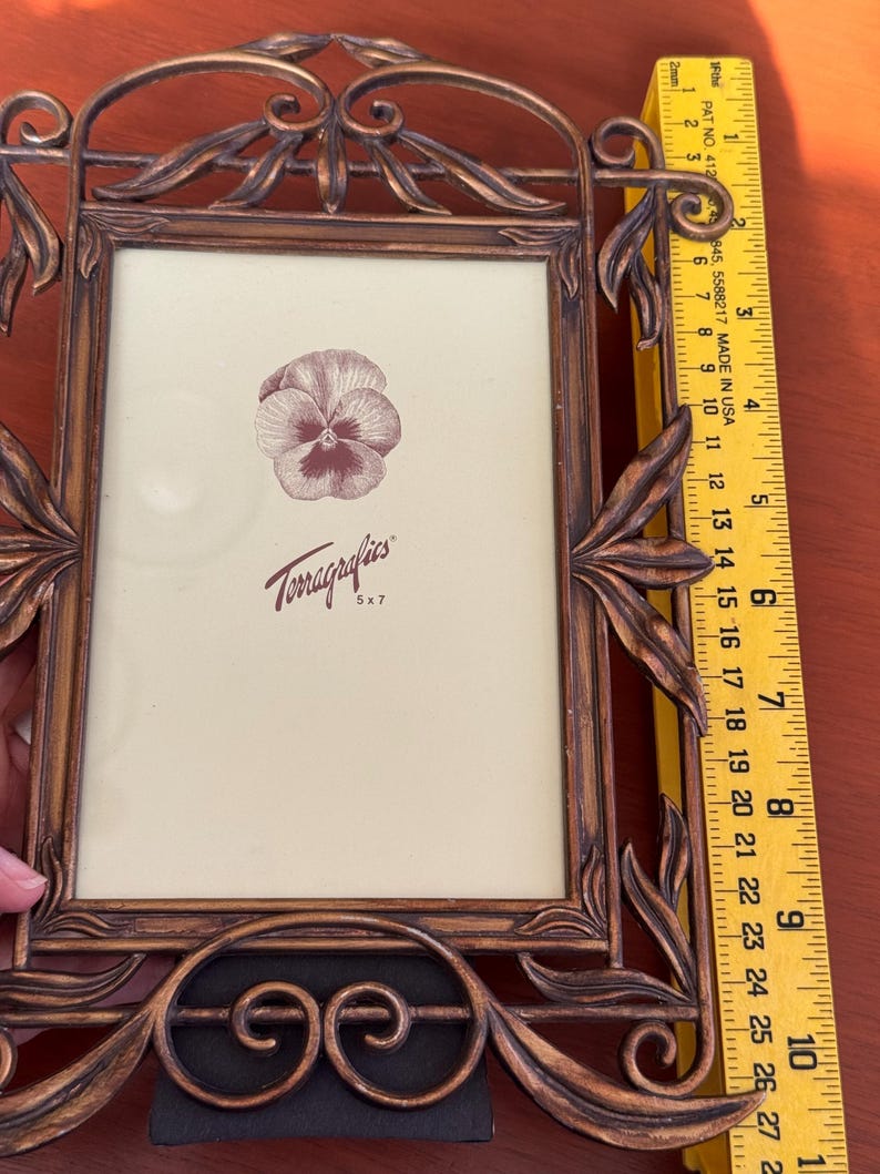 May include: A rectangular bronze-coloured metal picture frame with ornate leaf-like scrollwork detailing. A yellow ruler placed along the side indicates a height of approximately 25.4 cm. The frame contains a display card with a pansy illustration.