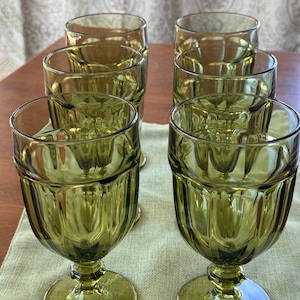 Op de afbeelding: Set van zes olijfgroene vintage waterglazen. De glazen hebben een textuur, geribbeld ontwerp op de kom en een ronde voet. De kelken zijn gerangschikt op een lichtgroene loper op een houten tafel.