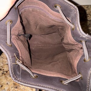 May include: A dark brown drawstring backpack with a brown interior. The bag has a textured exterior, metal eyelets, and drawstring closure. The interior features a zippered pocket.