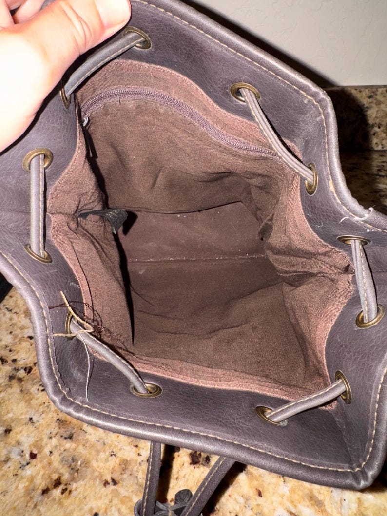 May include: A dark brown drawstring backpack with a brown interior. The bag has a textured exterior, metal eyelets, and drawstring closure. The interior features a zippered pocket.