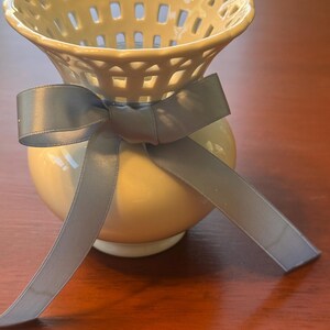 May include: A white ceramic vase featuring a flared, lattice-patterned rim and a smooth, rounded base. A light blue satin ribbon is tied in a bow around the neck of the vase, with two long ribbon ends hanging down the front of the ceramic body.