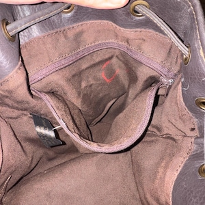May include: The interior of a dark brown bucket bag featuring a brown fabric lining, a zipped pocket, and brass-toned metal grommets along the top edge for a drawstring closure. A small black tag is attached to the interior side seam.