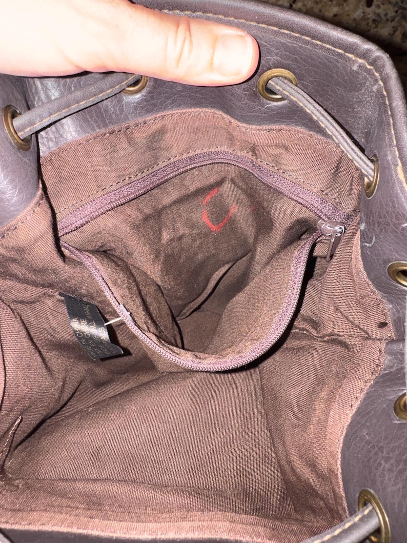 May include: The interior of a dark brown bucket bag featuring a brown fabric lining, a zipped pocket, and brass-toned metal grommets along the top edge for a drawstring closure. A small black tag is attached to the interior side seam.