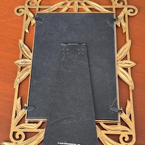 May include: A rectangular picture frame with an ornate gold-toned metal border featuring leaf motifs. The back is black with a hinged stand and a label reading 1996 TERRAGRAFICS, INC. MADE IN CHINA. The frame has four metal swivel clips.