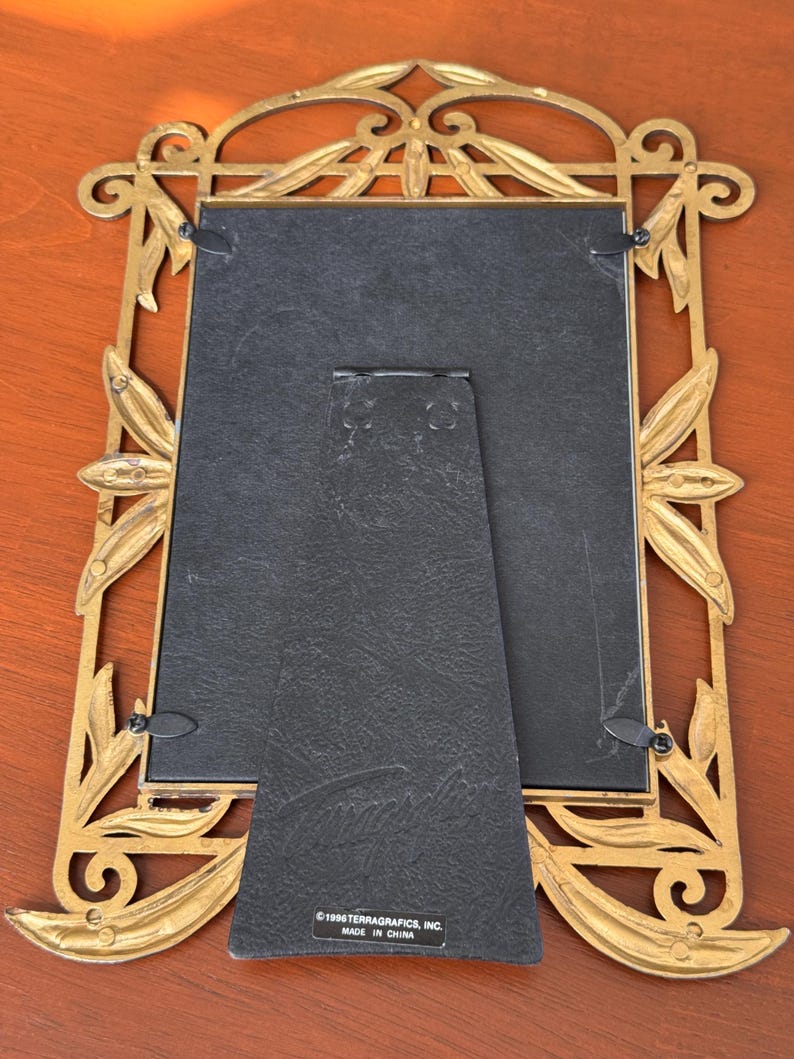 May include: A rectangular picture frame with an ornate gold-toned metal border featuring leaf motifs. The back is black with a hinged stand and a label reading 1996 TERRAGRAFICS, INC. MADE IN CHINA. The frame has four metal swivel clips.