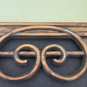 May include: A decorative metal object featuring a horizontal bar with two symmetrical, gold-toned scrollwork designs enclosed within an oval frame. The piece has a weathered, textured finish with visible signs of wear and sits against a dark background.