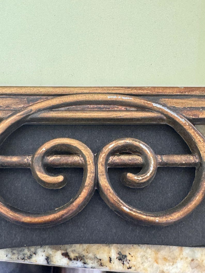 May include: A decorative metal object featuring a horizontal bar with two symmetrical, gold-toned scrollwork designs enclosed within an oval frame. The piece has a weathered, textured finish with visible signs of wear and sits against a dark background.