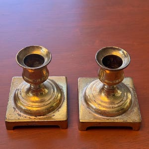 May include: A pair of matching brass candle holders featuring a square base and a circular top opening. The metal surface displays a polished gold-toned finish with visible signs of natural ageing and patina throughout the structure.