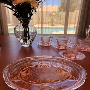 May include: A set of pink depression glass featuring a large oval serving platter and several matching teacups with saucers. Each piece displays a detailed pressed glass pattern with radiating fan motifs and intricate floral scrollwork designs.