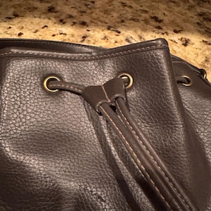 May include: A dark brown textured leather drawstring bag featuring brass-toned metal grommets and a matching leather drawstring cord. The bag has visible stitching along the edges and the drawstring closure mechanism is centred at the top.