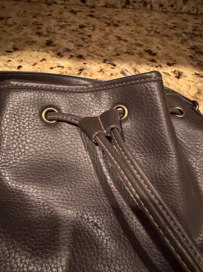 May include: A dark brown textured leather drawstring bag featuring brass-toned metal grommets and a matching leather drawstring cord. The bag has visible stitching along the edges and the drawstring closure mechanism is centred at the top.