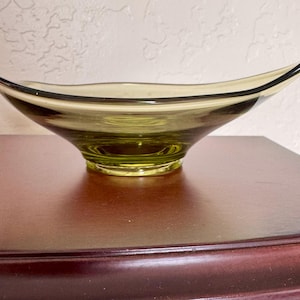 May include: A decorative, olive-green glass bowl with a flared, asymmetrical shape. The rim is outlined with a thin black band. The bowl sits on a small, circular base and is displayed on a dark brown surface.