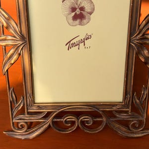 May include: A rectangular picture frame with a metallic bronze finish and ornate leaf-shaped accents along the sides and base. The cream-coloured insert features a brown pansy illustration and the text Terragraphics 5 x 7 in brown lettering.