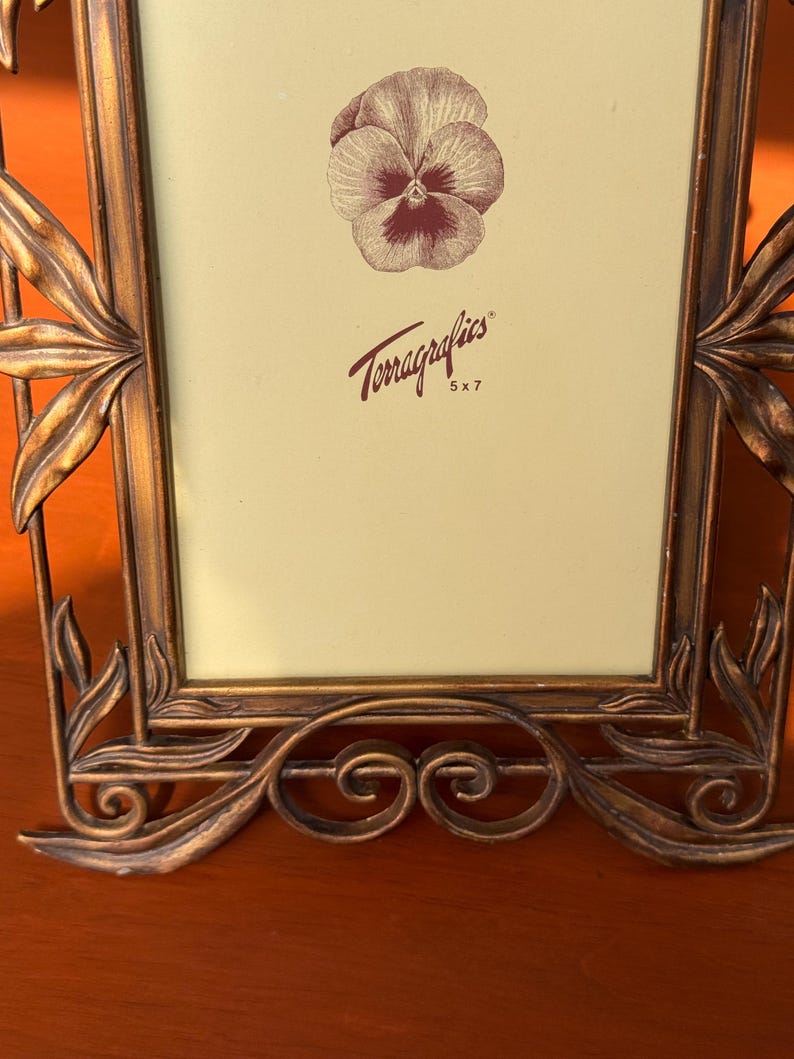 May include: A rectangular picture frame with a metallic bronze finish and ornate leaf-shaped accents along the sides and base. The cream-coloured insert features a brown pansy illustration and the text Terragraphics 5 x 7 in brown lettering.