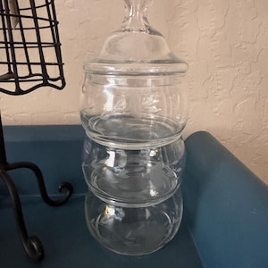 May include: A clear glass stackable canister set consisting of three rounded sections and a matching lid with a knob handle. Each glass section features a delicate etched floral vine pattern wrapping around the exterior surface.