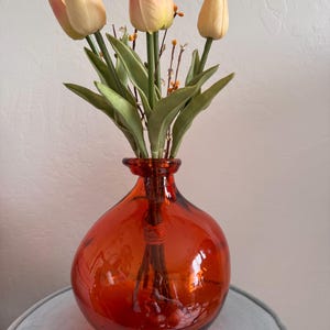 May include: An orange glass vase holds a bouquet of artificial tulips. The tulips have pale yellow petals with pink accents, green leaves, and small sprigs of berries. The vase has a rounded shape and sits on a light blue surface.