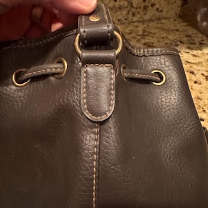 May include: Close-up of a dark brown leather handbag. The bag features gold-coloured metal hardware, including rings and eyelets. The stitching is visible, and the bag appears to be a bucket style.