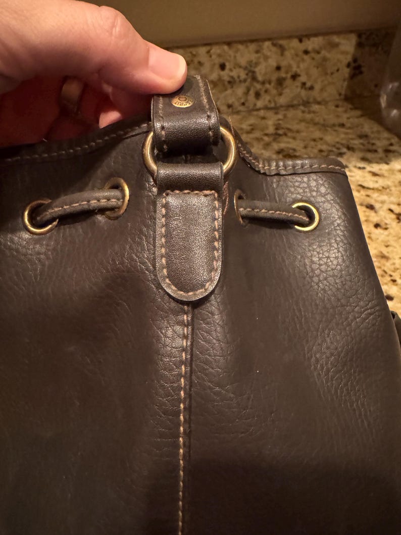 May include: Close-up of a dark brown leather handbag. The bag features gold-coloured metal hardware, including rings and eyelets. The stitching is visible, and the bag appears to be a bucket style.