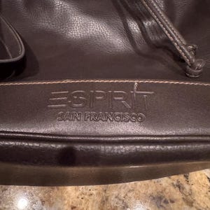 May include: A dark brown bucket bag with a drawstring closure and a textured surface. The front features the embossed text ESPRIT SAN FRANCISCO on a horizontal panel. The bag includes a shoulder strap and visible stitching details.