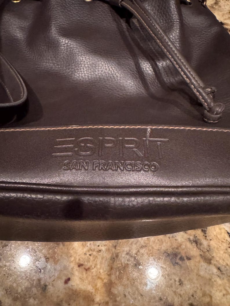 May include: A dark brown bucket bag with a drawstring closure and a textured surface. The front features the embossed text ESPRIT SAN FRANCISCO on a horizontal panel. The bag includes a shoulder strap and visible stitching details.