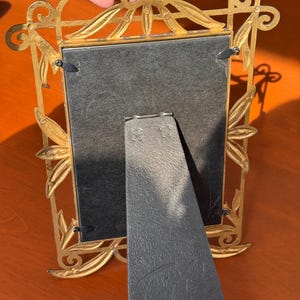 May include: A gold-coloured metal picture frame with an ornate leaf and vine design. The back features a black easel stand with a label that reads 1996 TERRAGRAFICS, INC. MADE IN CHINA. The frame is rectangular with four black metal swivel clips.