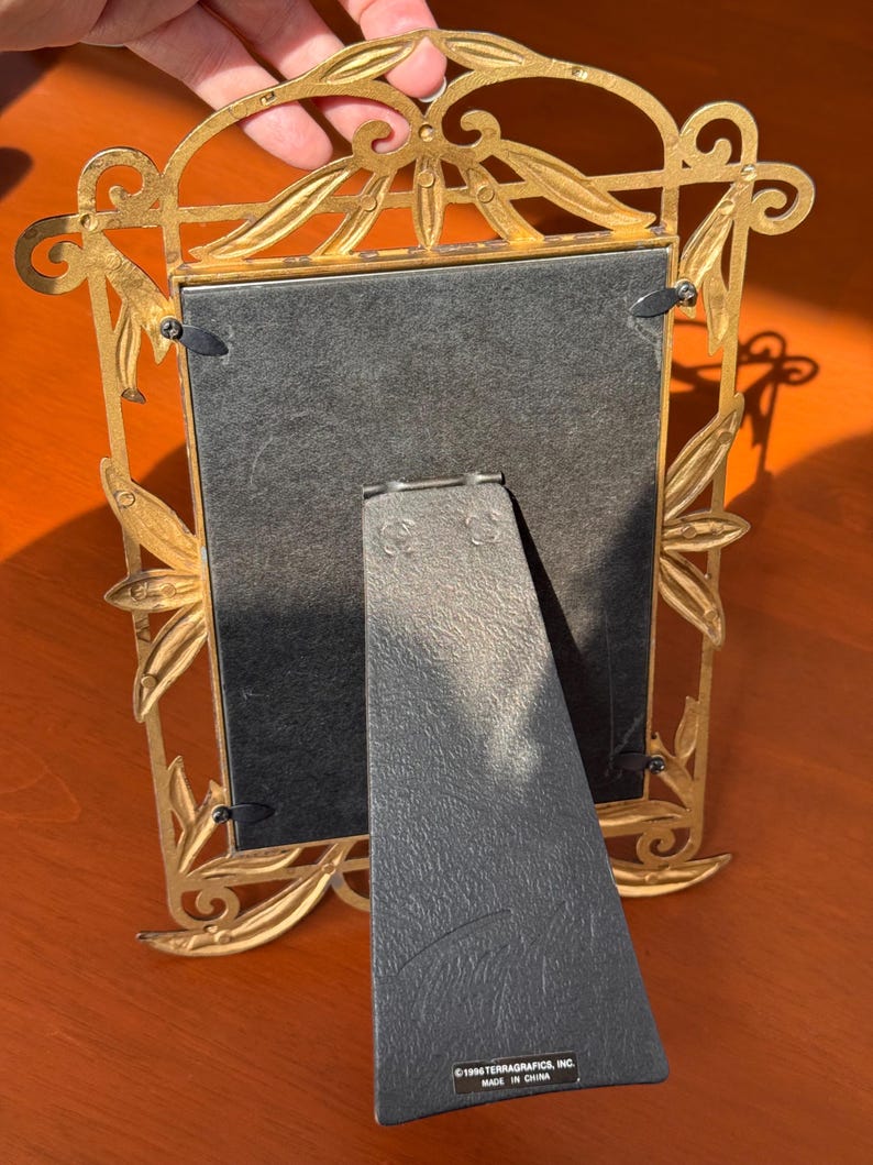 May include: A gold-coloured metal picture frame with an ornate leaf and vine design. The back features a black easel stand with a label that reads 1996 TERRAGRAFICS, INC. MADE IN CHINA. The frame is rectangular with four black metal swivel clips.