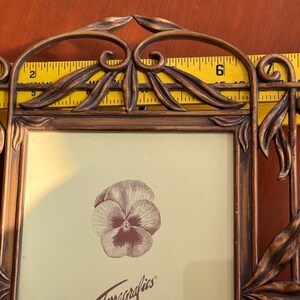 May include: A rectangular bronze-toned metal picture frame featuring decorative leaf and scrollwork accents. A yellow measuring tape is positioned behind the frame, showing a width of approximately 20 cm. A pansy illustration is inside.