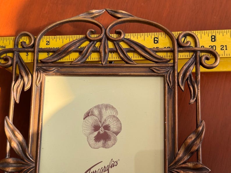 May include: A rectangular bronze-toned metal picture frame featuring decorative leaf and scrollwork accents. A yellow measuring tape is positioned behind the frame, showing a width of approximately 20 cm. A pansy illustration is inside.