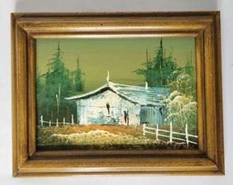 May include: A framed oil painting depicting a white farmhouse with a green roof, surrounded by trees and a white picket fence. The painting has a rustic and charming feel.