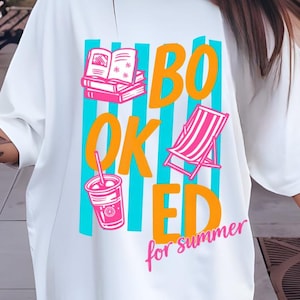 May include: White t-shirt with a colorful graphic that reads "BOOKED for summer." The design includes illustrations of books, a beach chair, and a drink. The text and illustrations are in shades of orange, pink, and blue.