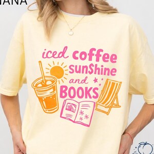May include: A light yellow t-shirt with the words "iced coffee sunshine and BOOKS" in pink and orange. The graphic includes an iced coffee, sun, beach chair, and an open book. The word "BANANA" is printed in black at the top left.