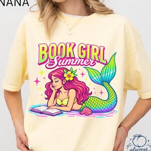 May include: A light yellow t-shirt with the text "BOOK GIRL Summer" above a mermaid illustration. The mermaid has pink hair, a yellow bikini, and a colorful tail. The shirt also has the text "BANANA" at the top left.