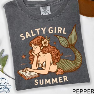 May include: A dark gray t-shirt with a mermaid graphic. The illustration features a mermaid with red hair, a flower, and a green tail, reading a book. The words "Salty Girl Summer" are printed above and below the mermaid.