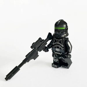 Könnte beinhalten: Eine schwarze Minifigur mit grünem Visier und weißen Akzenten, die ein schwarzes Gewehr hält. Die Figur ist ein Spielzeug, wahrscheinlich ein Sammlerstück. Der Hintergrund ist weiß.