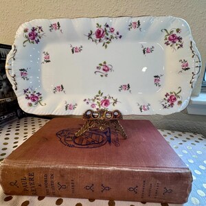 May include: A rectangular white porcelain serving tray featuring a delicate floral pattern of pink roses and small purple flowers. The tray has a scalloped edge with gold trim detailing along the rim and handles on both ends.