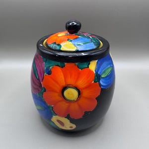 May include: A black ceramic jar with a lid, adorned with vibrant floral designs. The jar features a large orange flower with a yellow center, surrounded by blue, purple, and yellow flowers. The lid is also decorated with colorful flowers.