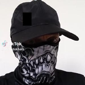 May include: A person wearing a black cap and a monster-themed face mask. The mask features a detailed, grayscale design of a monster's face. The text "monster facemask" is visible at the top of the image.