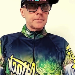 Peut inclure: Un homme portant une casquette noire et des lunettes, et une chemise de pêche. La chemise est bleu marine avec un motif coloré et les mots "Fish Attack" en blanc. La chemise est en microfibre, tissu léger.