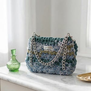 May include: A handmade, blue and green crocheted handbag with silver chain straps. The bag has a rectangular shape and features a label that reads "Água de Sol". A green glass bottle and a gold tray with rings are also visible.