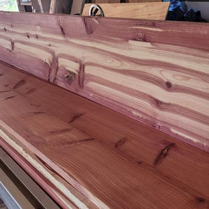 May include: Close-up of freshly cut cedar wood planks, displaying rich reddish-brown hues with lighter, cream-coloured streaks. The wood grain is prominent, revealing natural patterns. The planks are stacked, ready for woodworking projects.