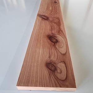Puede incluir: Una tabla rectangular de madera de color marrón rojizo, que muestra un patrón de vetas natural con varios nudos. La madera tiene una superficie texturizada y probablemente está destinada a la carpintería o la artesanía. Las dimensiones de la tabla son en centímetros.
