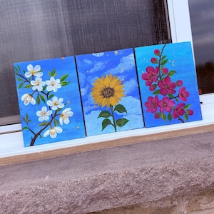 May include: Three square paintings featuring floral designs. One shows white blossoms on a branch, another a yellow sunflower, and the third, pink flowers. All are set against a blue background with gold accents, creating a cohesive floral art set.