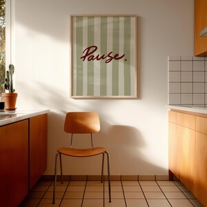 May include: A framed wall art print featuring vertical sage green and white stripes with the word Pause. written in a dark red cursive script. The print is displayed in a light wood frame hanging on a plain white wall above a wooden chair.