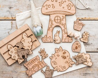 May include: Wooden playset with a mushroom house, a squirrel, a bunny, a beehive, a flower, a watering can, a welcome sign, and a tree with apples. The pieces are on a white fabric surface.