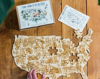 May include: A wooden jigsaw puzzle of the USA with a map outline. The puzzle pieces are decorated with illustrations of animals and plants native to the United States. The puzzle is partially assembled, with some pieces missing. The box for the puzzle is visible in the background, with the title "Flora - Fauna of the USA Puzzle".