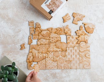 May include: A wooden Oregon puzzle with detailed laser-cut designs of the state's regions. The puzzle pieces are scattered on a light surface, with a hand placing a piece. The box reads "OREGON PUZZLE".