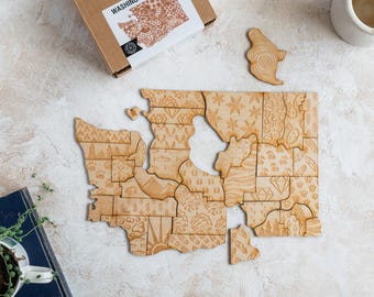 May include: A wooden jigsaw puzzle of the state of Washington, with each county represented by a different pattern. The puzzle is laid out on a light beige surface, with the box for the puzzle in the top left corner. The box is brown with the words "WASHINGTON PUZZLE" printed on it.