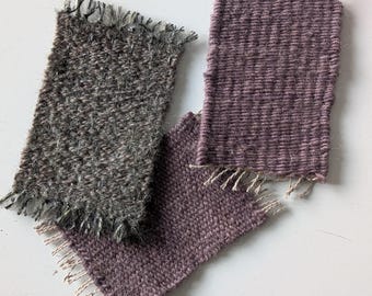 May include: Three rectangular woven fabric swatches with fringed edges. One swatch is a dark charcoal gray with a textured, fuzzy appearance. The other two swatches are a muted mauve purple color, featuring a distinct horizontal woven pattern.