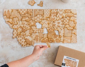 May include: A wooden jigsaw puzzle of the state of Montana, with each county represented by a different pattern. The puzzle is laid out on a light beige surface, with a hand placing a piece into the puzzle. A brown box with the text "MONTANA PUZZLE" is in the lower right corner of the image.